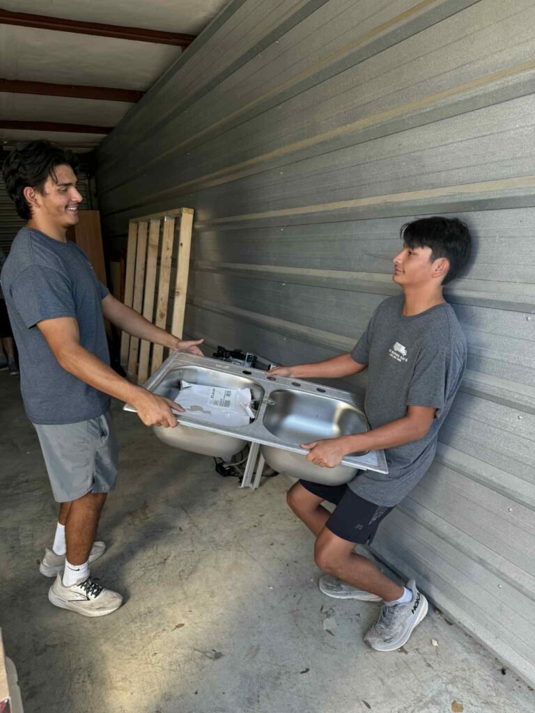 Two professional movers from The Moving Guys carefully carrying a stainless steel double kitchen sink inside a moving truck, preparing it for safe transport.